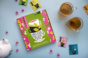 Box of Clipper Tea on light blue background surrounded by small pink flowers and tea sachets.